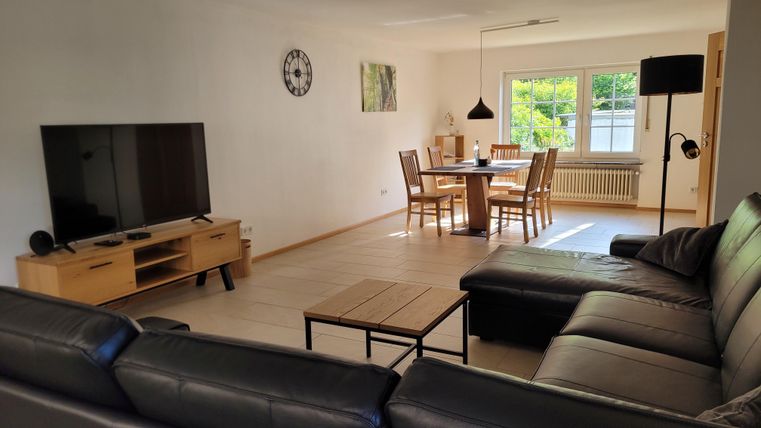 A modern living room with a large TV, a comfortable couch, and a dining area. It has many windows that let in natural light.