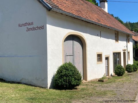 Une maison d'art avec une façade blanche et un toit rouge. L'entrée est décorée d'une grande porte en bois et de petits buissons.