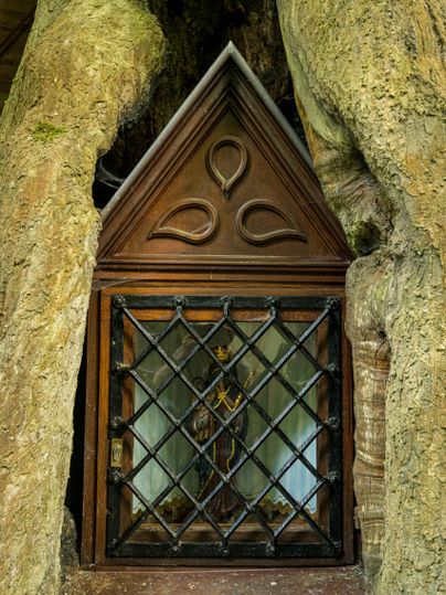 A small, wooden-decorated church is embedded between two thick tree trunks. It has a pointed roof and a barred door.