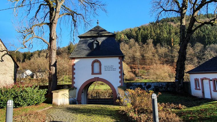 Ein historisches Torhaus mit einem Bogen, umgeben von Bäumen und sanften Hügeln. Der Himmel ist klar und blau.