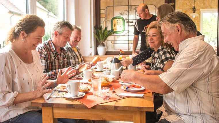 A group of five people is sitting together at a table and enjoying breakfast. On the table are cups, plates with food, and fresh snacks.