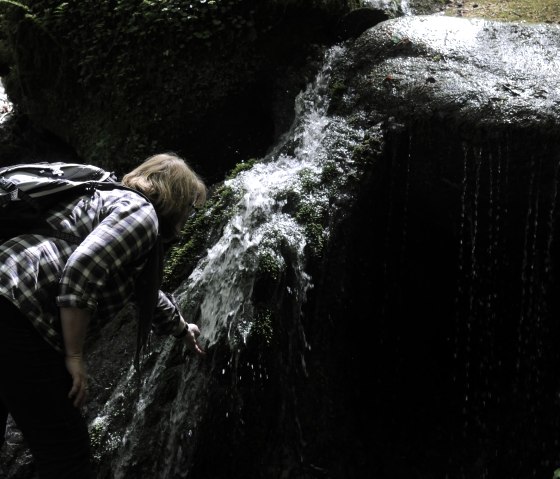 Une personne portant un sac &agrave; dos se penche sur une petite chute d'eau recouverte de mousse dans la for&ecirc;t., &copy; Felsenland S&uuml;deifel Tourismus GmbH
