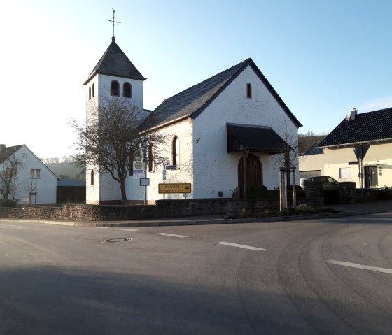 Eine kleine Kirche mit Turm in Wi&szlig;mannsdorf, umgeben von Wohnh&auml;usern, bei tiefstehender Sonne. Ein Wegweiser zeigt Richtung Arzfeld und Koosb&uuml;sch., &copy; Georg Lotzkes