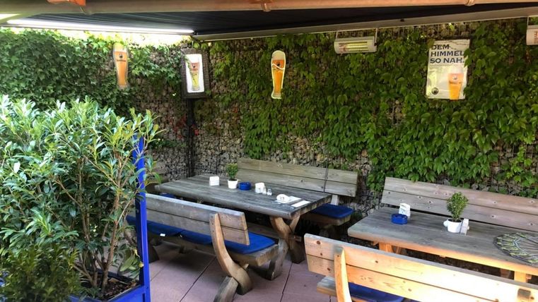 A cozy outdoor area with wooden tables and benches. The walls are decorated with plants and beer posters are hanging.