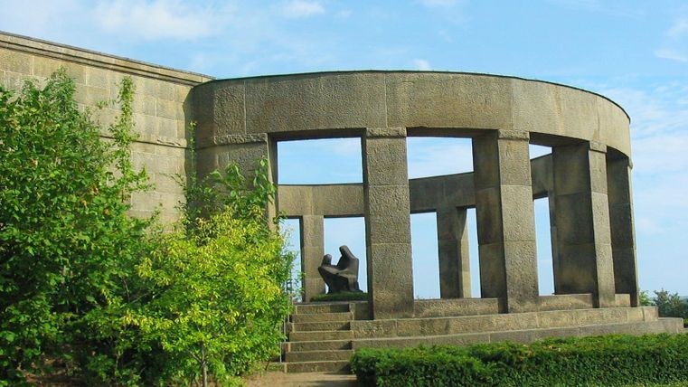 Ein historisches Denkmal mit runden Steinbögen und einer Skulptur im Inneren. Umgeben von grünen Pflanzen und einem blauen Himmel.