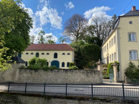 A historic building with light walls surrounded by green plants. In the background, there are trees and a blue sky.