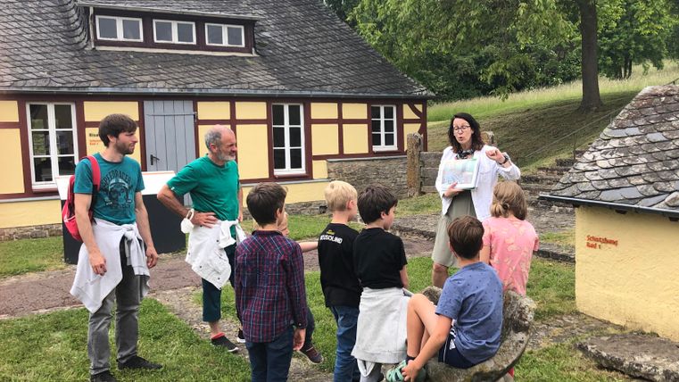 A group of children is listening to a woman who is explaining something. In the background, a historical building can be seen in a green environment.
