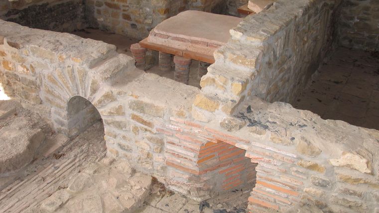 Eine archäologische Stätte mit alten Steinmauern und Überresten von Gebäuden. Die Mauern zeigen frühere Bauweisen und Strukturen.