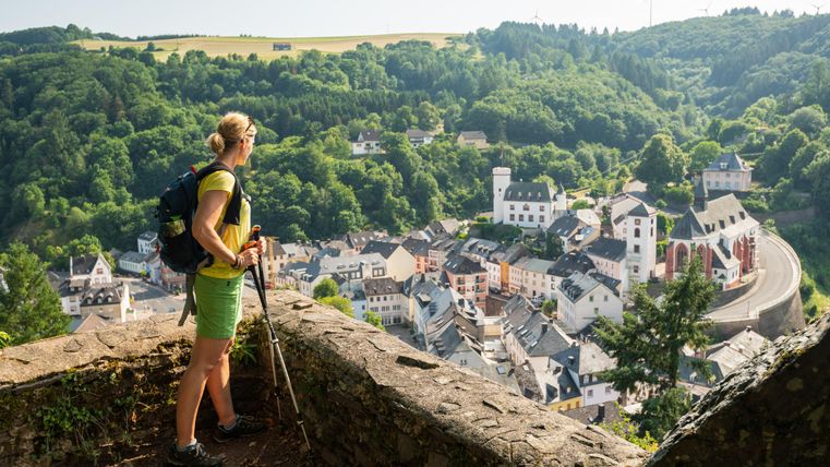 Une randonneuse se tient à un point de vue et regarde un village pittoresque au milieu de collines verdoyantes. La scène est calme et montre la beauté naturelle des environs.