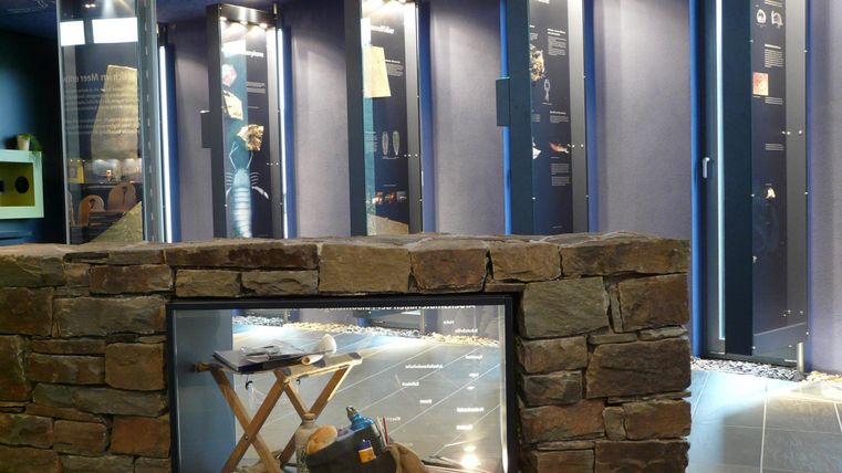 An exhibition room with several display cases showcasing historical objects. In the foreground, a stone pedestal with a table and various collectibles is visible.