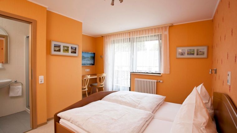 A cozy room with orange walls and a double bed. There is a television, a table with chairs, and a window that lets in plenty of light.