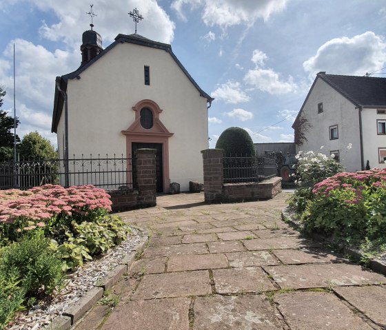 Dorfkirche in Sefferweich, umgeben von bl&uuml;henden Beeten und einem gepflasterten Weg, unter einem blauen Himmel mit Wolken., &copy; Tourist-Information Bitburger Land
