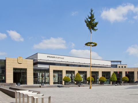 A modern building with large windows and a clear sky. In the foreground, trees and a paved area can be seen.