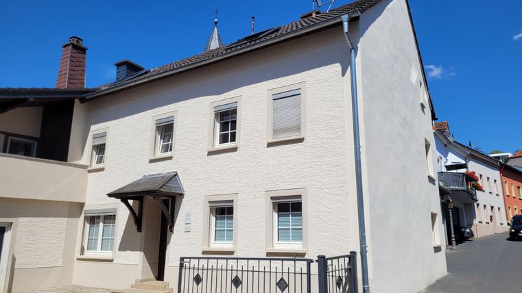 A modern, bright house with a white facade. Next to the house, a paved road leads with adjacent residential buildings.