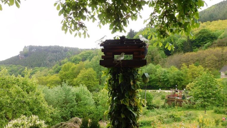 A tranquil garden with lush greenery and a small birdhouse. Gentle hills can be seen in the background.
