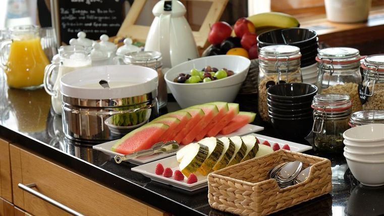 A beautifully arranged breakfast buffet with fresh fruit, including watermelons, melons, and grapes. In the background, various glasses and containers stand for more delicacies.