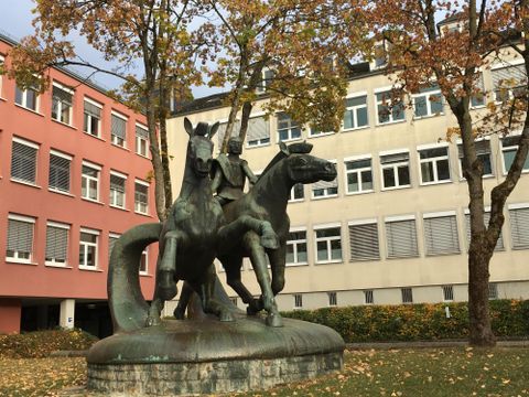 A bronze sculpture of two horses with a rider is located in an urban area. In the background, modern buildings and trees with autumn leaves can be seen.