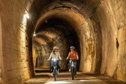 Zwei Radfahrerinnen fahren durch einen beleuchteten Tunnel. Die Wände sind aus Backstein und die Atmosphäre ist ruhig und geheimnisvoll.