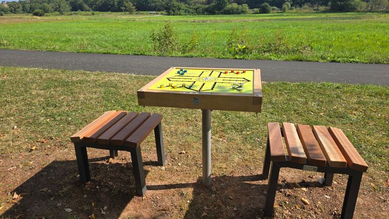 A table with a game board and two benches stands in a meadow. In the background, you can see green fields and a blue sky.