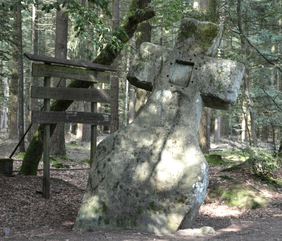Een groot, met mos begroeid stenen kruis staat in het bos naast een houten wegwijzer. Zonlicht schijnt door de bomen., &copy; Lauschtour.de
