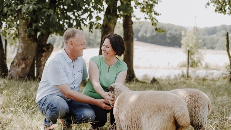 A smiling couple is kneeling in the greenery and petting sheep. In the background, trees and a gentle landscape can be seen.