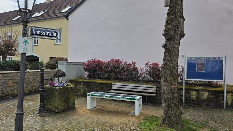 Een rustige hoek met een bank en een straatnaambord "Nimstraat". Op de achtergrond zijn bloemen en een boom zichtbaar, omringd door klinkers.