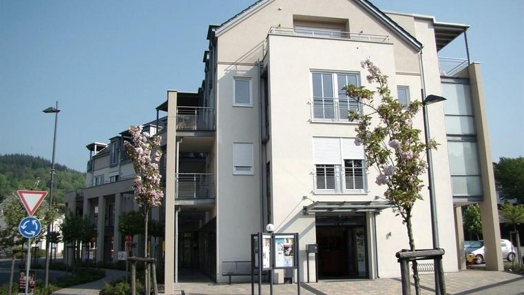 A modern building with clean lines and large windows. Planted areas and a small street are visible in front of the building.