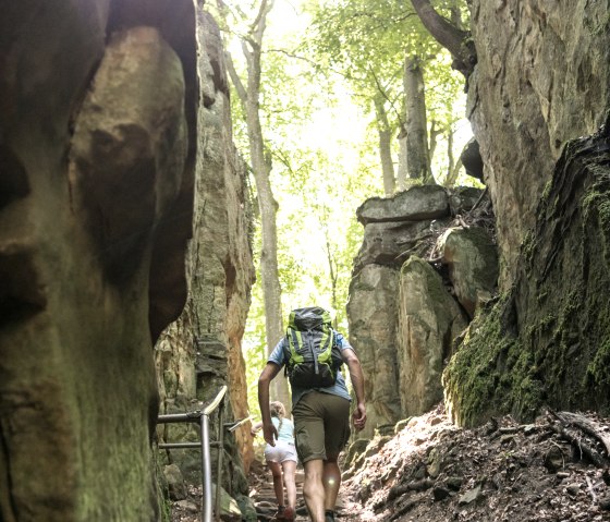 Zwei Personen wandern durch die Teufelsschlucht, umgeben von hohen Felsen und B&auml;umen. Sonnenlicht strahlt durch das Bl&auml;tterdach., &copy; Eifel Tourismus GmbH, Dominik Ketz