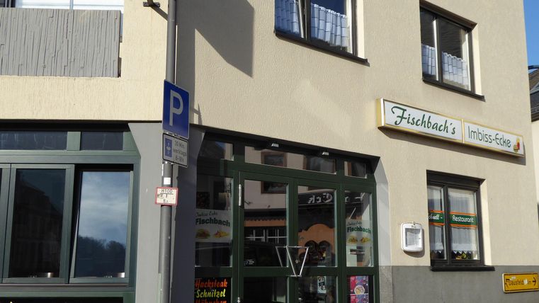 Een snackbar met de naam "Fischbach's" in een licht gebouw. De ramen zijn groot en er is een zonnescherm en een parkeerbord voor de deur.