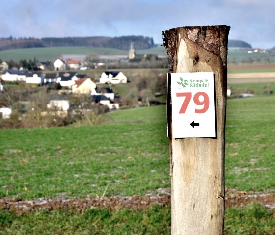 Un poteau en bois avec un panneau du parcours n&deg; 79 du parc naturel de l'Eifel du Sud, avec en arri&egrave;re-plan un paysage rural avec des maisons et des champs., &copy; TI Bitburger Land