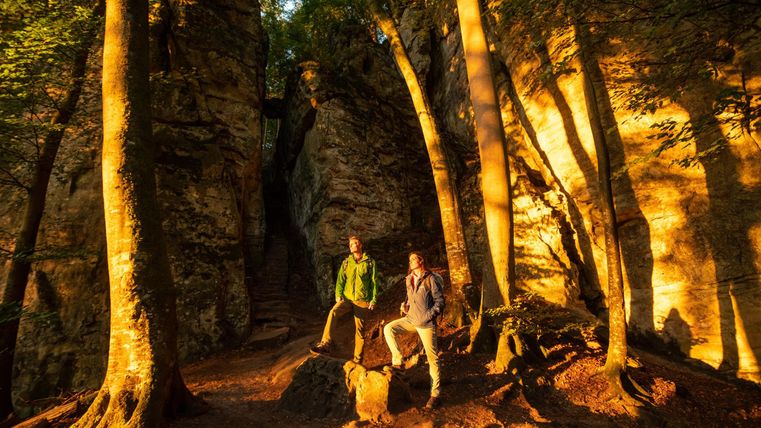 Zwei Personen stehen in einem Wald zwischen hohen Felsen. Das Sonnenlicht wirkt warm und beleuchtet die Umgebung.