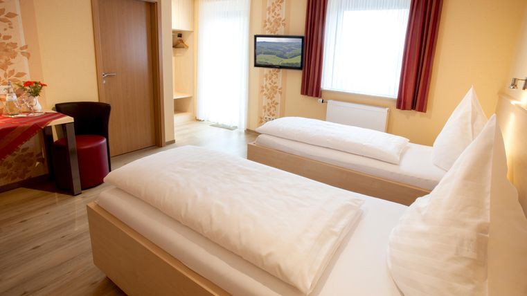 A cozy hotel room with two single beds and a table. The room is bright and cheerful with windows that let in daylight.