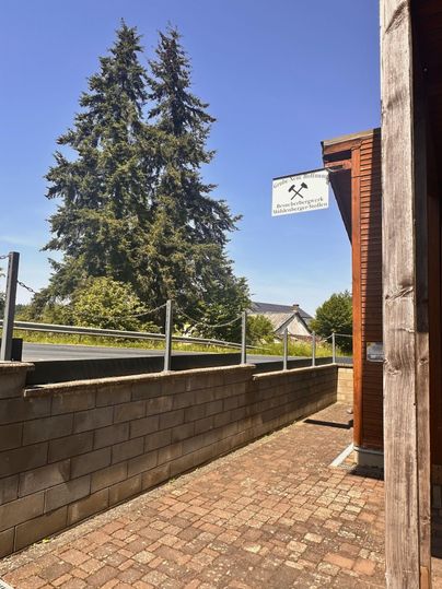 Une rue étroite avec un trottoir en pavés et un mur en pierres. À l'arrière-plan, on aperçoit de grands arbres et un panneau.