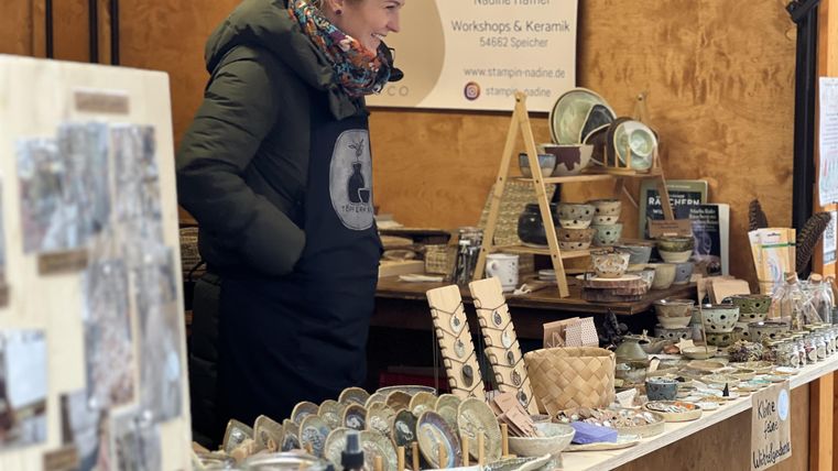 Een verkoopster staat aan een tafel vol handgemaakte keramiek. Op de achtergrond is een bord te zien met informatie over workshops en de aangeboden producten.