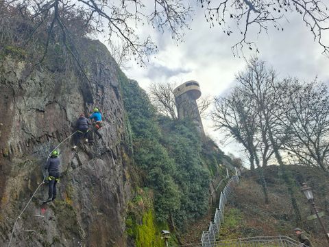 Deux grimpeurs escaladent une paroi rocheuse, tandis qu'en arrière-plan une tour d'observation est visible. Des arbres entourent la scène et le ciel est nuageux.