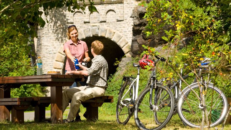 Zwei Personen machen eine Pause an einem Picknicktisch neben Fahrrädern, im Hintergrund eine Steinbrücke.