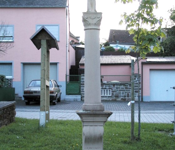 Une croix de chemin en pierre se trouve sur la place d'un village, entour&eacute;e d'herbe. En arri&egrave;re-plan, on aper&ccedil;oit un arbre et une maison rose., &copy; Conny Meier