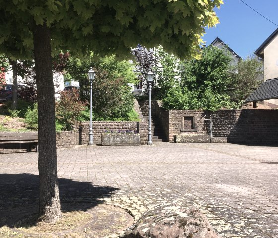 Une place de village pav&eacute;e &agrave; Gransdorf avec un grand arbre, des lanternes et une grosse pierre au premier plan. Le ciel est clair et bleu., &copy; Tourist-Information Bitburger Land