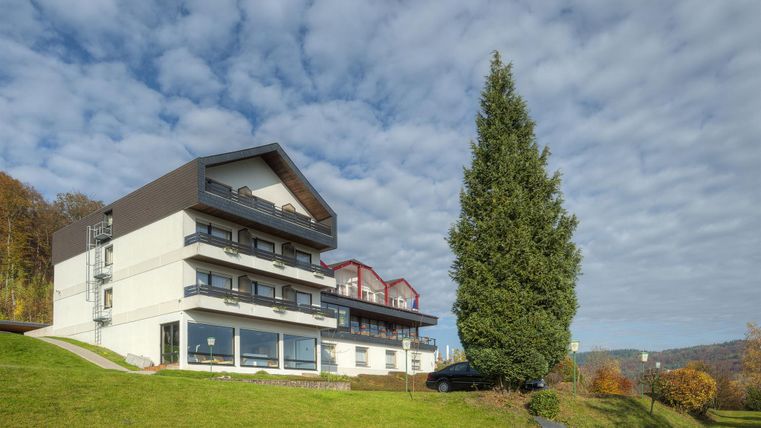 Ein modernes Gebäude mit mehreren Etagen in einer grünen Umgebung. Der Himmel ist bewölkt und die Landschaft ist hügelig.