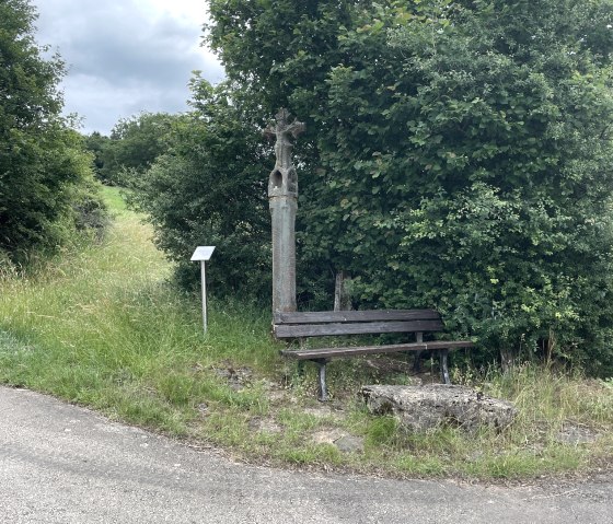 Een wegkruis en een bankje staan aan de rand van een pad, omringd door groene struiken en bomen., &copy; Daniel K&ouml;hler