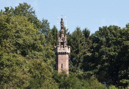 Die Mariens&auml;ule in Kyllburg ragt aus dichtem Wald hervor. Eine Statue steht auf einem steinernen Turm, umgeben von gr&uuml;nen B&auml;umen und blauem Himmel., &copy; TI Bitburger Land