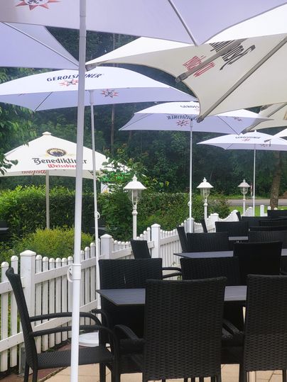 A cozy terrace with black chairs and tables under white umbrellas. In the background, green bushes and a white fence are visible.