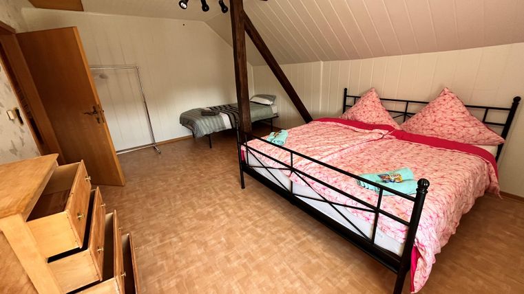 Een gezellige slaapkamer met een tweepersoonsbed en roze beddengoed. Naast het bed staat een commode en er is een extra slaapplaats in de hoek.