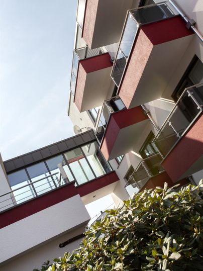A modern residential building with several balconies and large windows. The façade is designed in light and red tones.