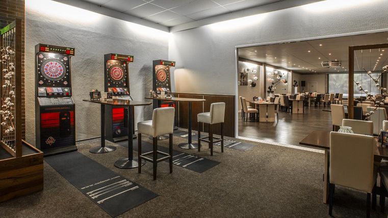 Een modern restaurant met een dartgebied en lichte tafels. De ruimte is vriendelijk ingericht en biedt plaats aan gasten.