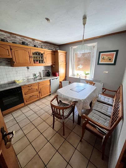 Een lichte keuken met houten kasten en een grote eettafel. Er zijn gezellige stoelen en ramen met daglicht.