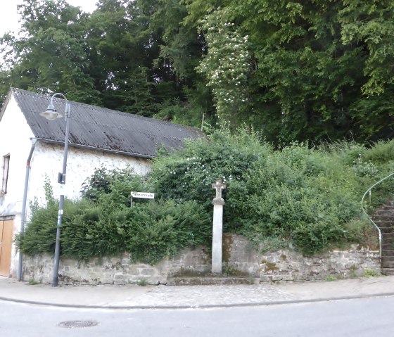 Ancien b&acirc;timent au coin d'une rue avec croix et escaliers, entour&eacute; de verdure. Panneau routier 'R&ouml;merstra&szlig;e' visible., &copy; Elke Wagner, Felsenland S&uuml;deifel Tourismus GmbH