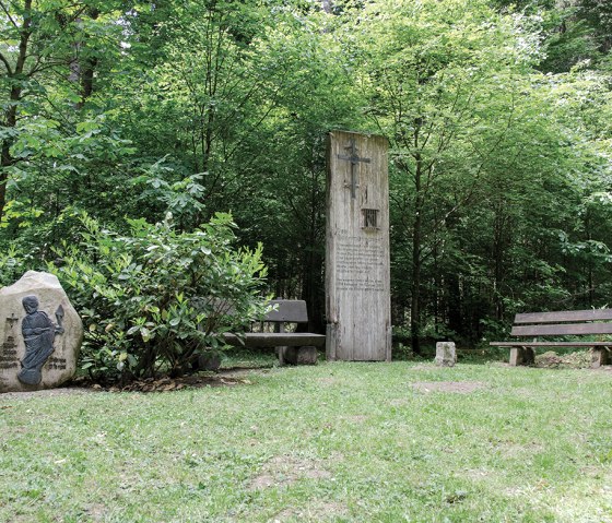 Ein Holzdenkmal und ein Steinrelief im Wald, umgeben von B&auml;nken und B&auml;umen. Die Szene wirkt ruhig und einladend., &copy; TI Bitburger Land