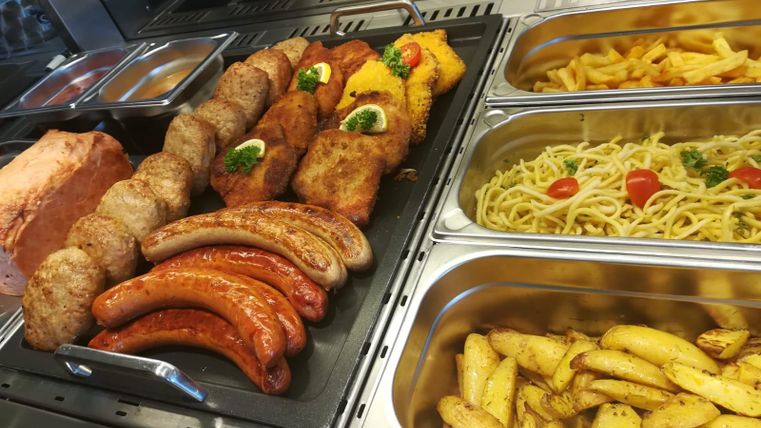Une sélection de différents plats de viande, y compris des saucisses et des galettes, est présentée dans un comptoir. À côté des types de viande, il y a aussi des pâtes et des quartiers de pommes de terre.