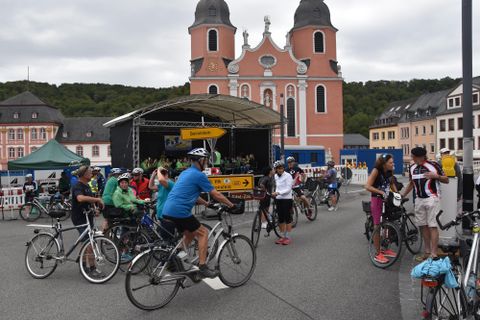 Een groep fietsers verzamelt zich voor een historische kerk. Op de achtergrond zijn gebouwen en een podium te zien.